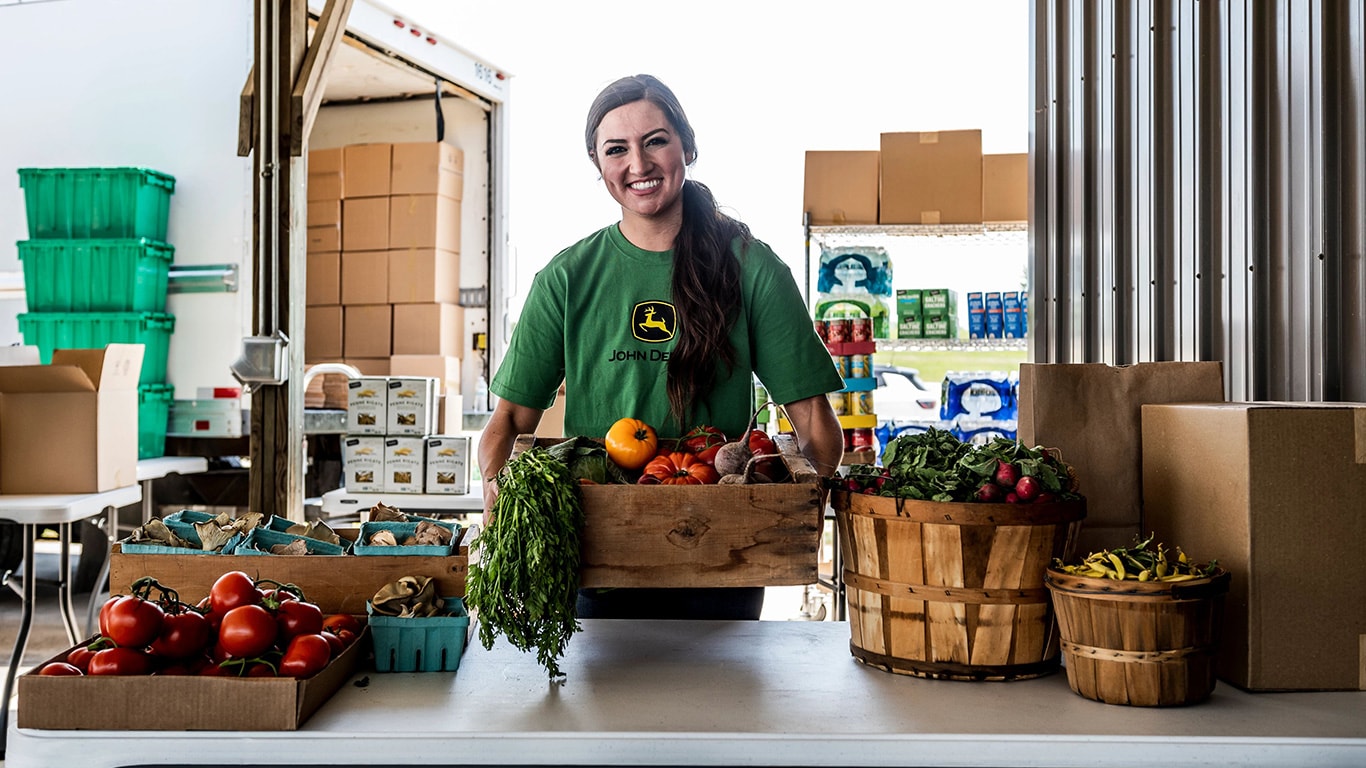 A woman in a John Deere shirt with a many fresh fruit in baskets in front of her.