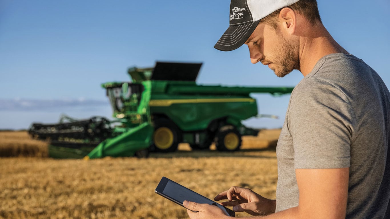 Man looking at a tablet in a field in front of a John Deere combine
