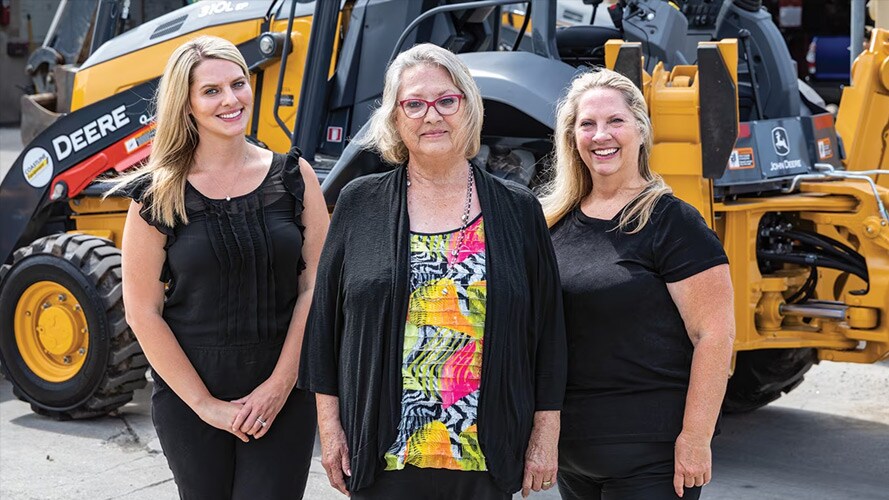 Amanda Matson, Janet Ferguson et Nikki Worden de Newman Backhoe Service se tiennent devant une rétrocaveuse 310L de John Deere
