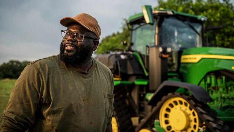 Un fermier noir se tenant fièrement près d'un tracteur John Deere