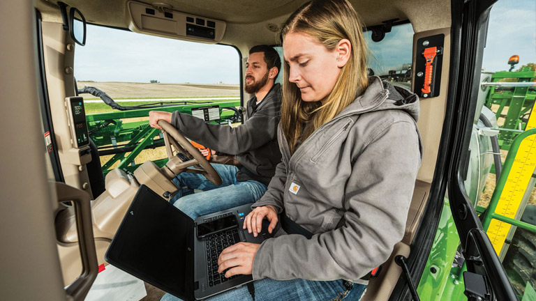 A man and a woman in a John Deere tractor cab inputting data into a laptop