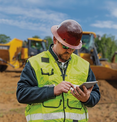 Un travailleur de la construction portant un gilet de sécurité vert regarde son appareil mobile en avant d'une chargeuse et d'un camion-benne.