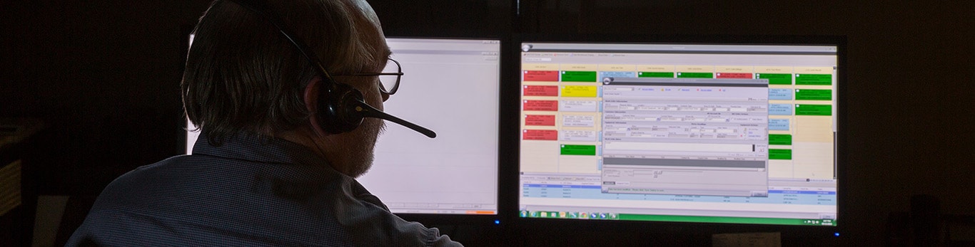 An analyst looking at data on two computer screens and talking on a headset in a dark room.