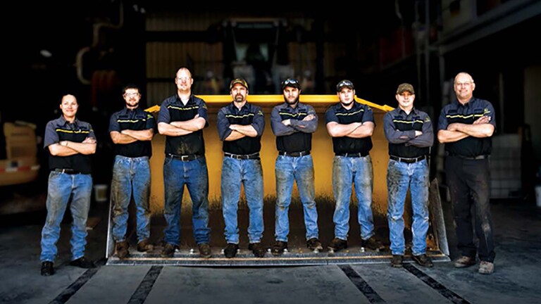 multiple mechanics standing in front of a large yellow plow