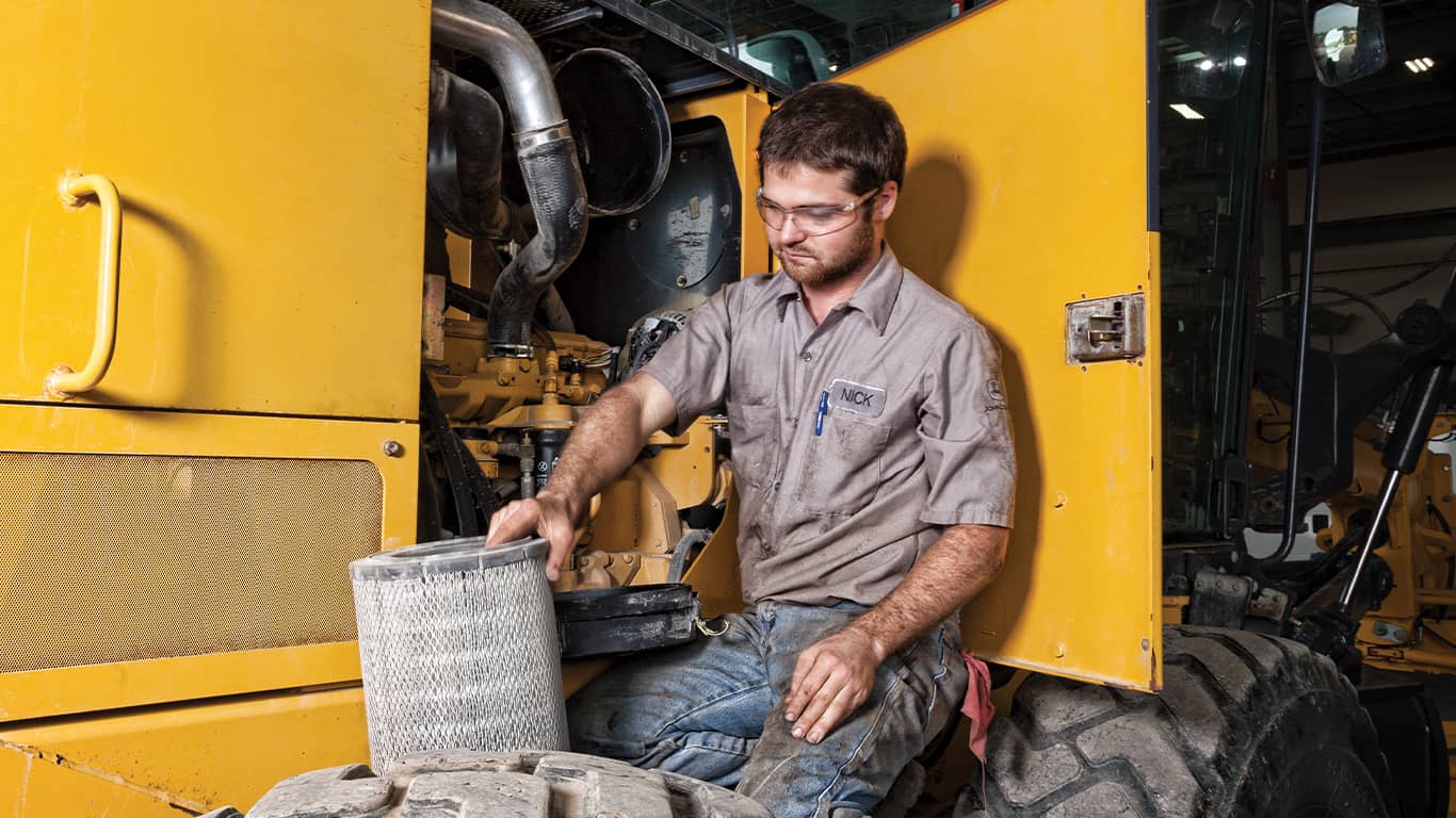 Un homme portant des lunettes de protection et travaillant sur un moteur John Deere à l’intérieur d’un équipement lourd