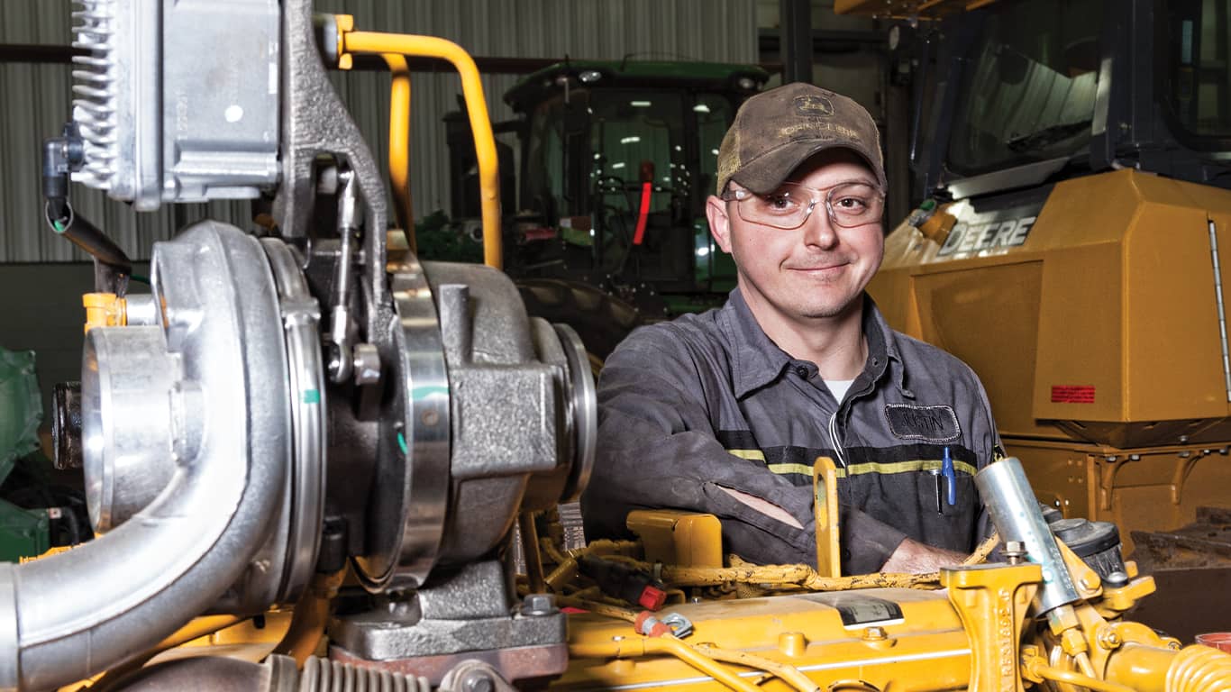 Un homme portant des lunettes de protection sourit à côté d’un moteur John Deere