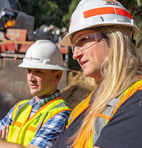 Male and female workers wearing construction hats and safety vests looking at a piece of construction equipment