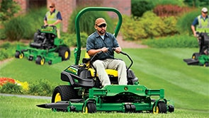 Three people operating commercial mowers in a grassy field with well manicured flora in the background.