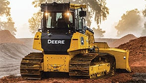 John Deere construction machine pushing dirt on a job site with trees in the background.