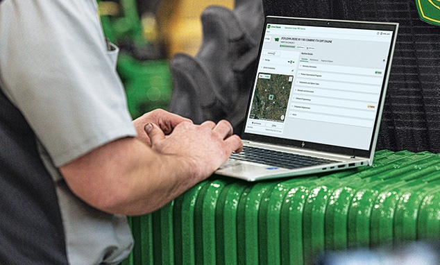 Person using a laptop displaying John Deere Operations Center tools while seated on equipment.