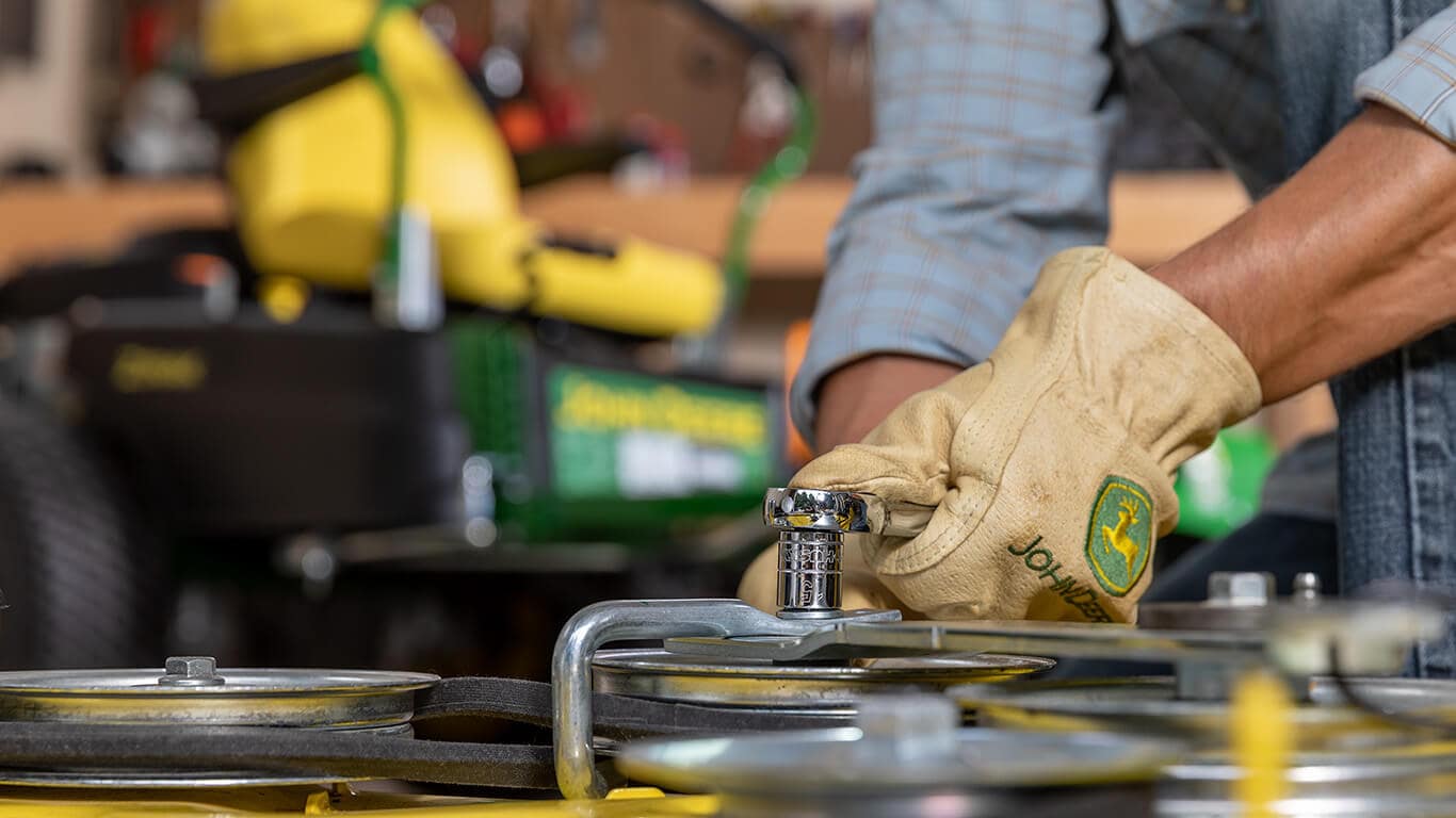Close-up on a person's hands in John Deere branded gloves wrenching on a machine with a blurry zero-turn mower in the background.