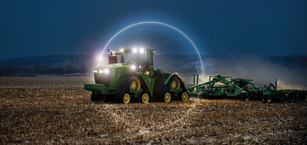 Large tracked tractor pulling tillage equipment across a field at night with work lights on.