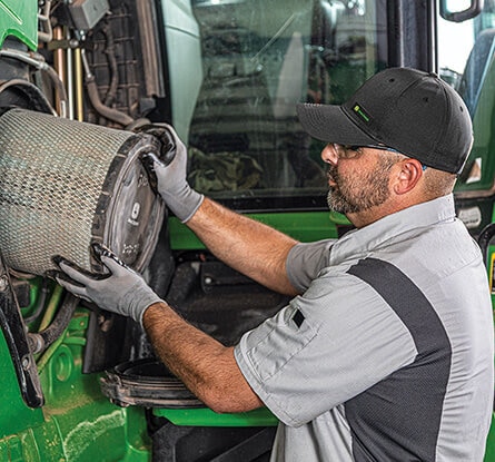 Person installing a large cylindrical air filter into the engine compartment of a green tractor.