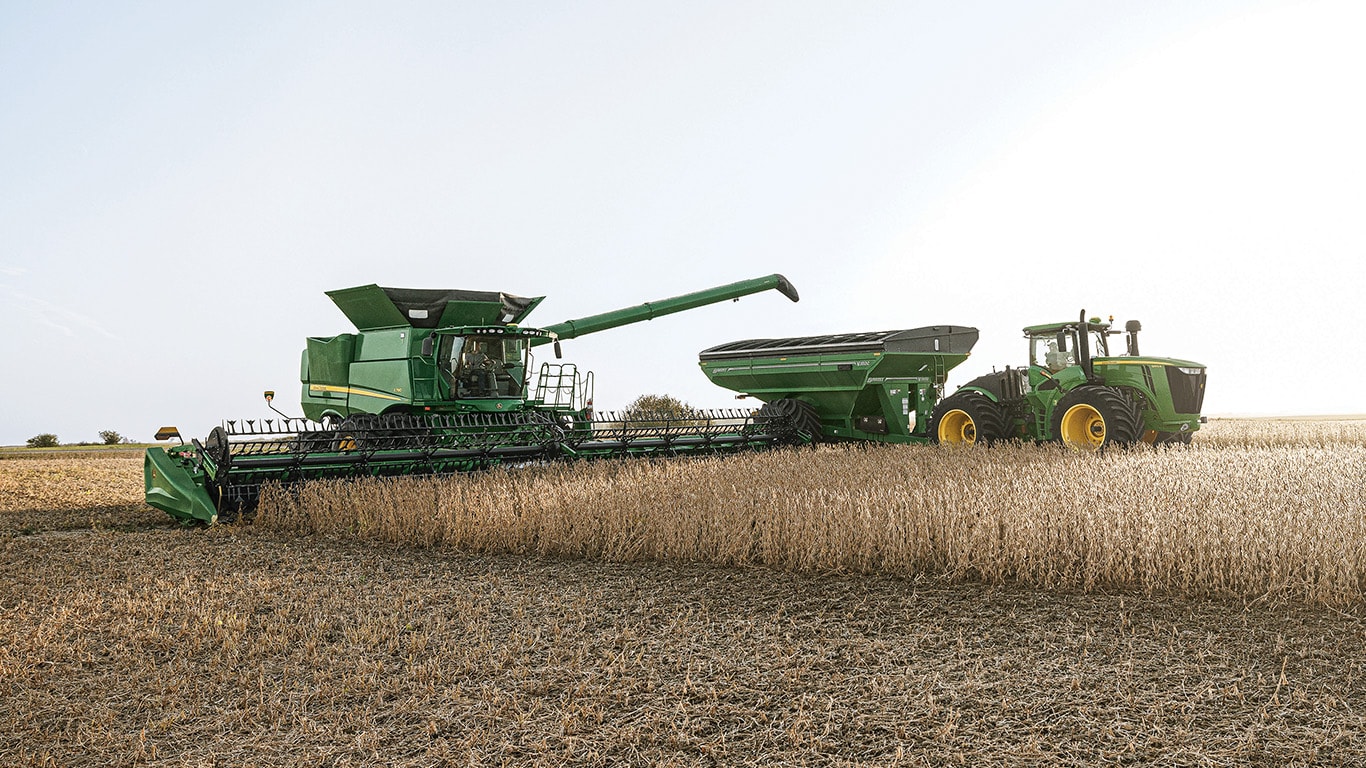 John&nbsp;Deere combine harvesting a grain field while unloading into a grain cart pulled by a tractor, with cut and uncut crop visible under a clear sky.
