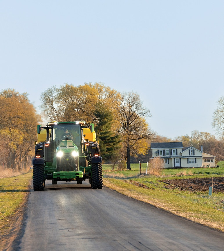 Green John Deere tractor driving on a rural road near a farmhouse and agricultural fields.