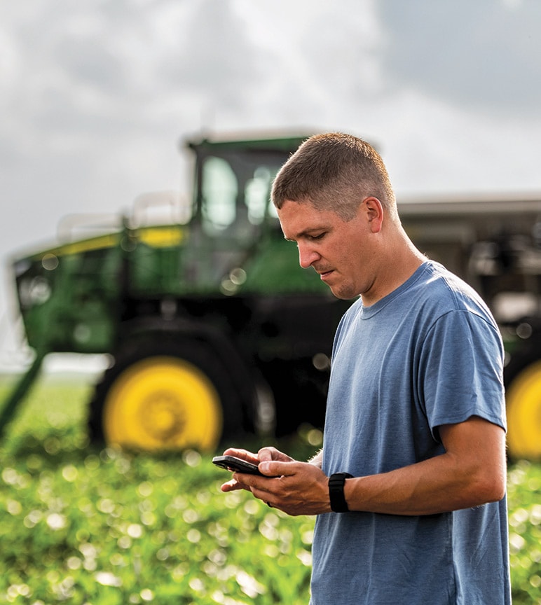 Person using a smartphone in front of John Deere equipment while reviewing warranty information.