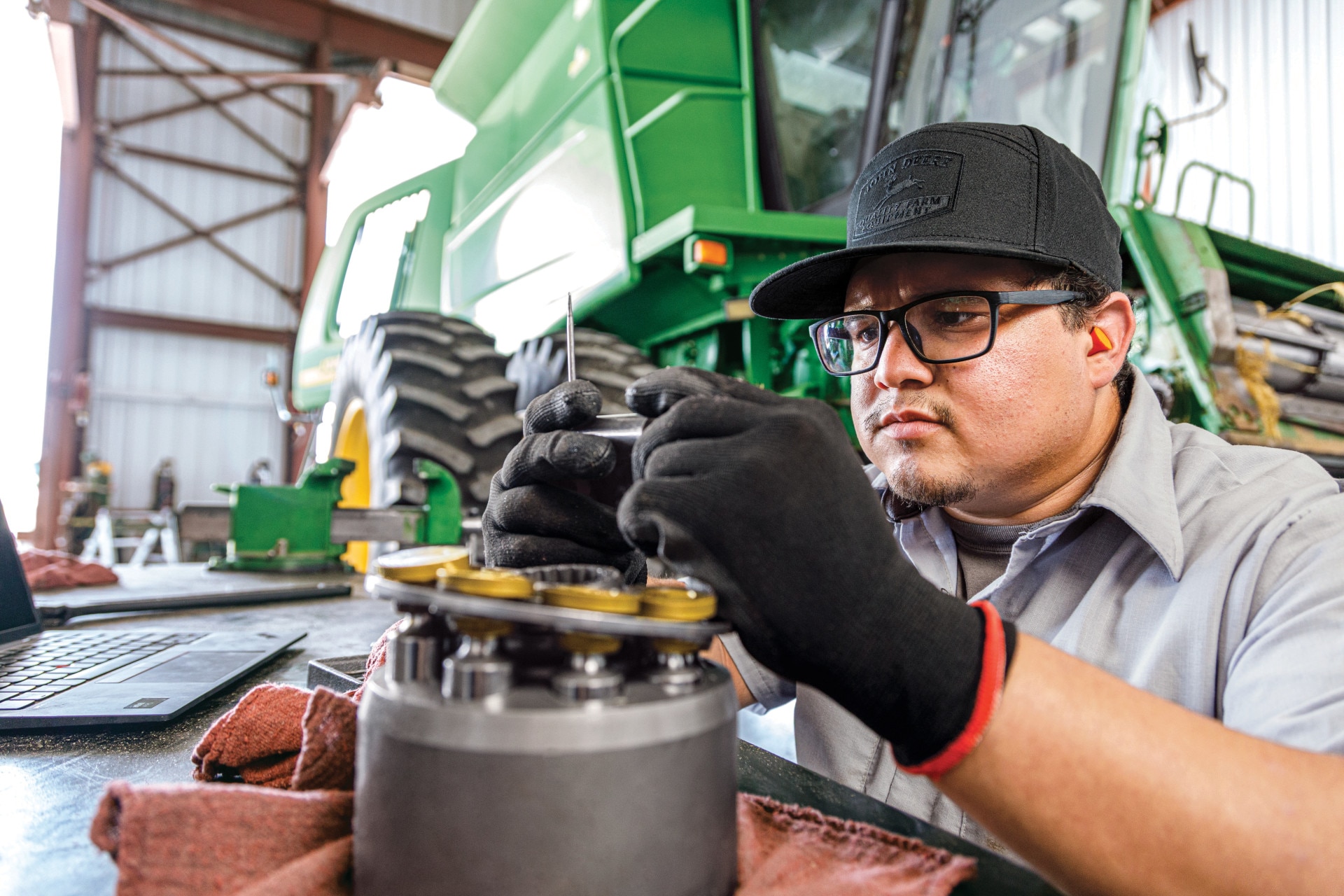 Technician assembling or servicing a mechanical component while working beside a John Deere machine in a workshop.