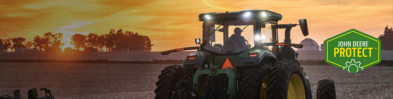 Rear view of a green John Deere tractor working in a field at sunrise or sunset, with illuminated cab lights and agricultural equipment visible in the foreground.