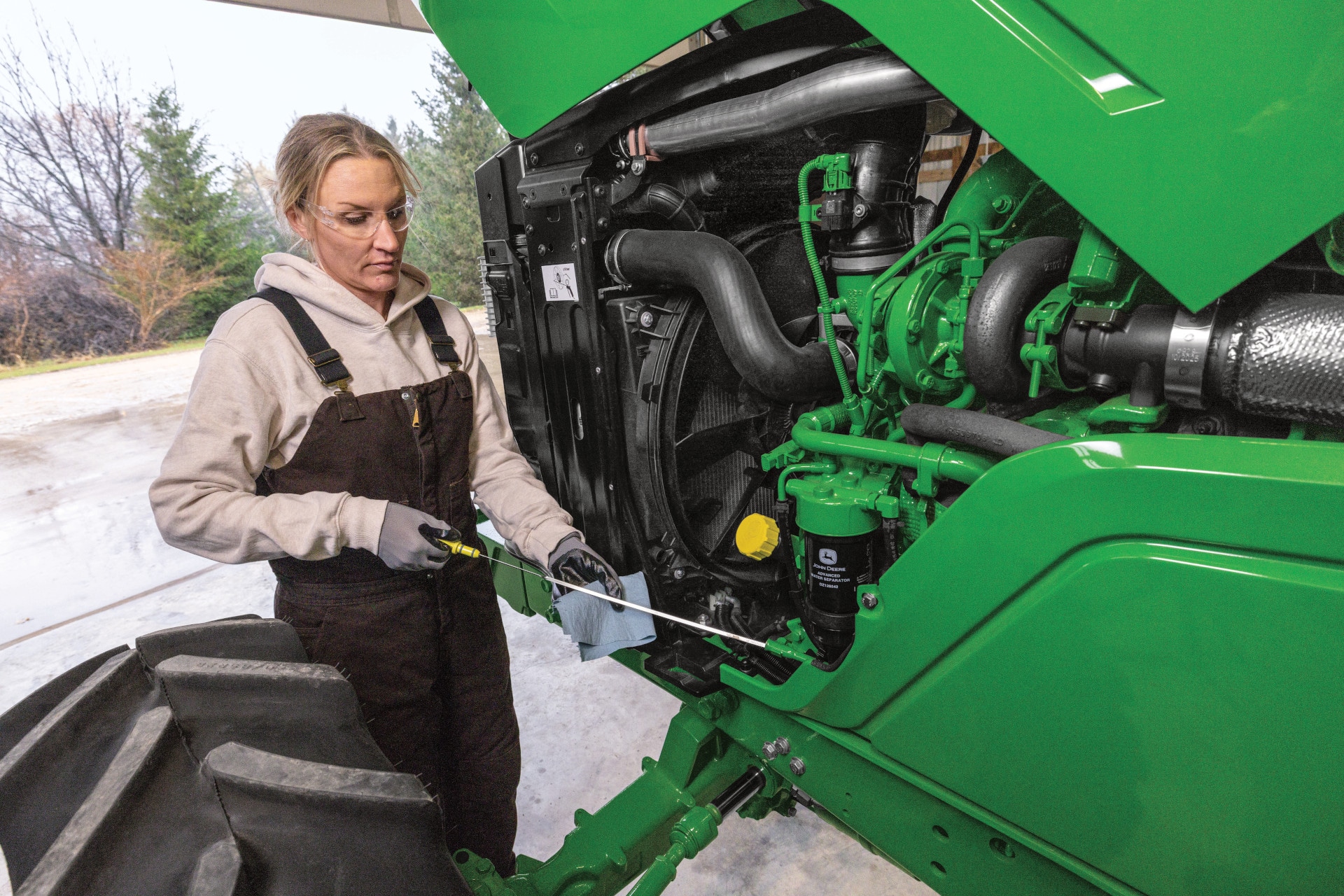 Person performing maintenance on an open John Deere machine compartment, inspecting engine components with a hand tool.