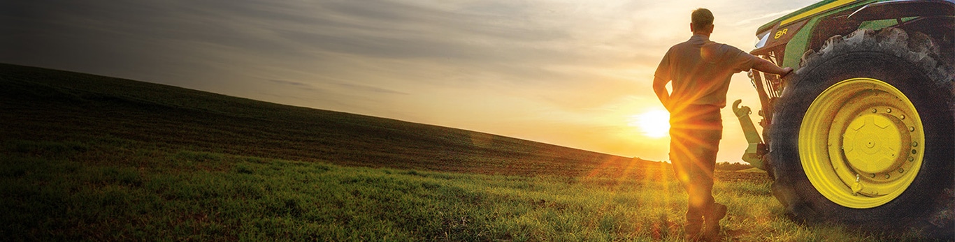 Man standing with his arm on an 8R Tractor tire looking out towards the sunset.
