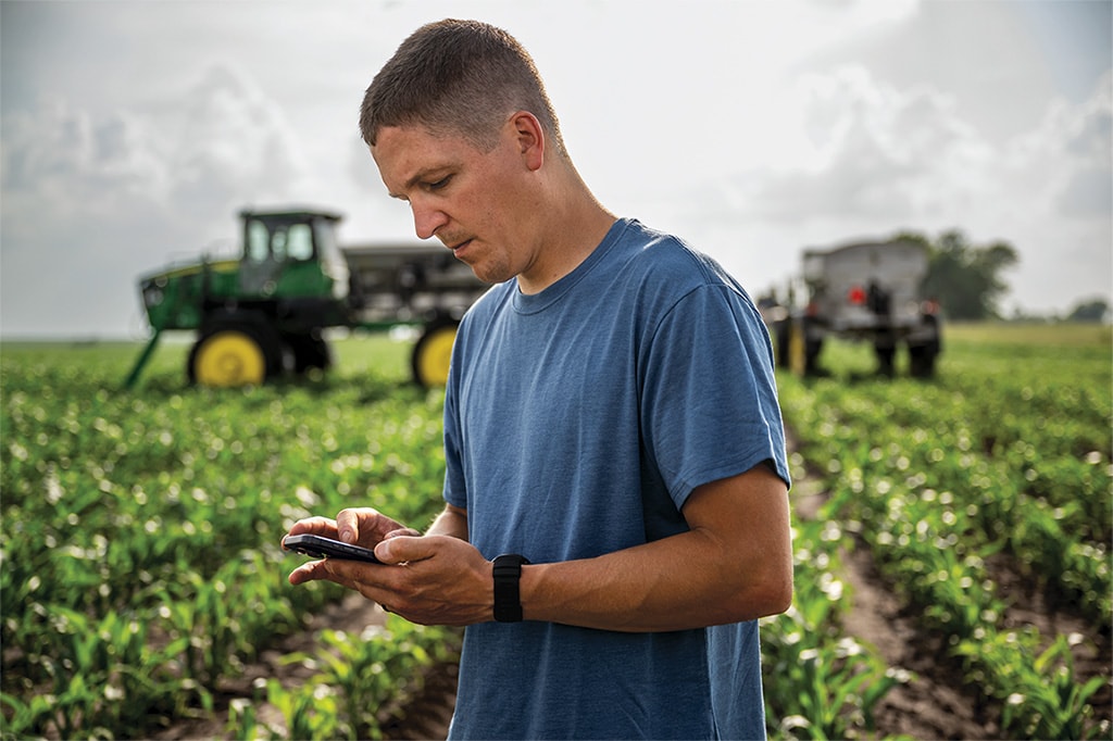 Homme dans un champ regardant son téléphone avec un équipement agricole au loin.