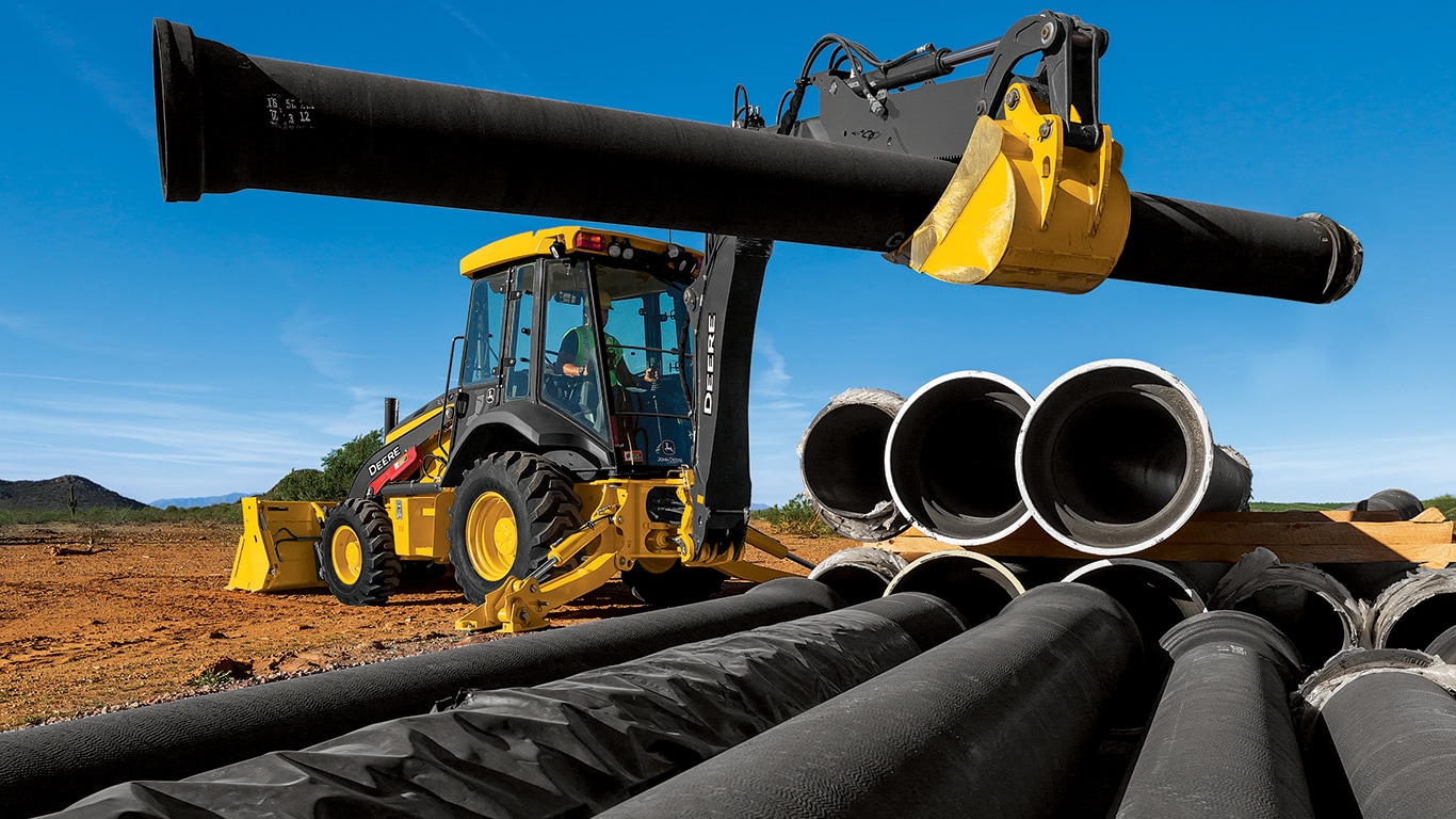A 310G Backhoe using the boom arm to move large black pipes.