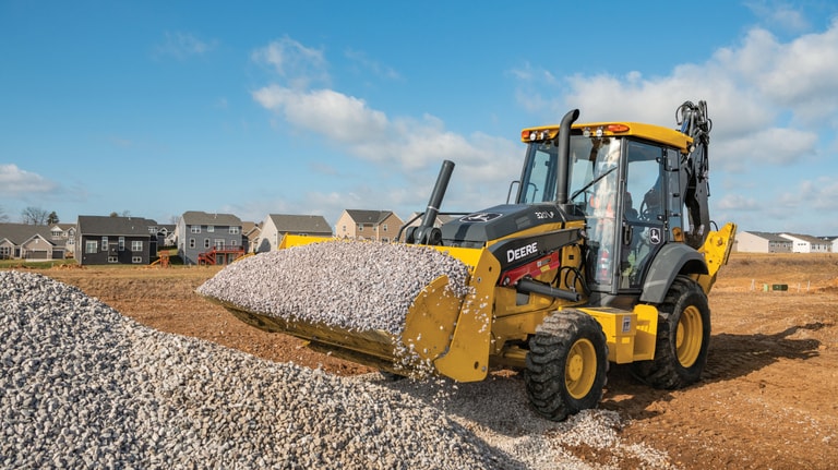 A 320P Backhoe hauling small rocks in the bucket with houses in the background.