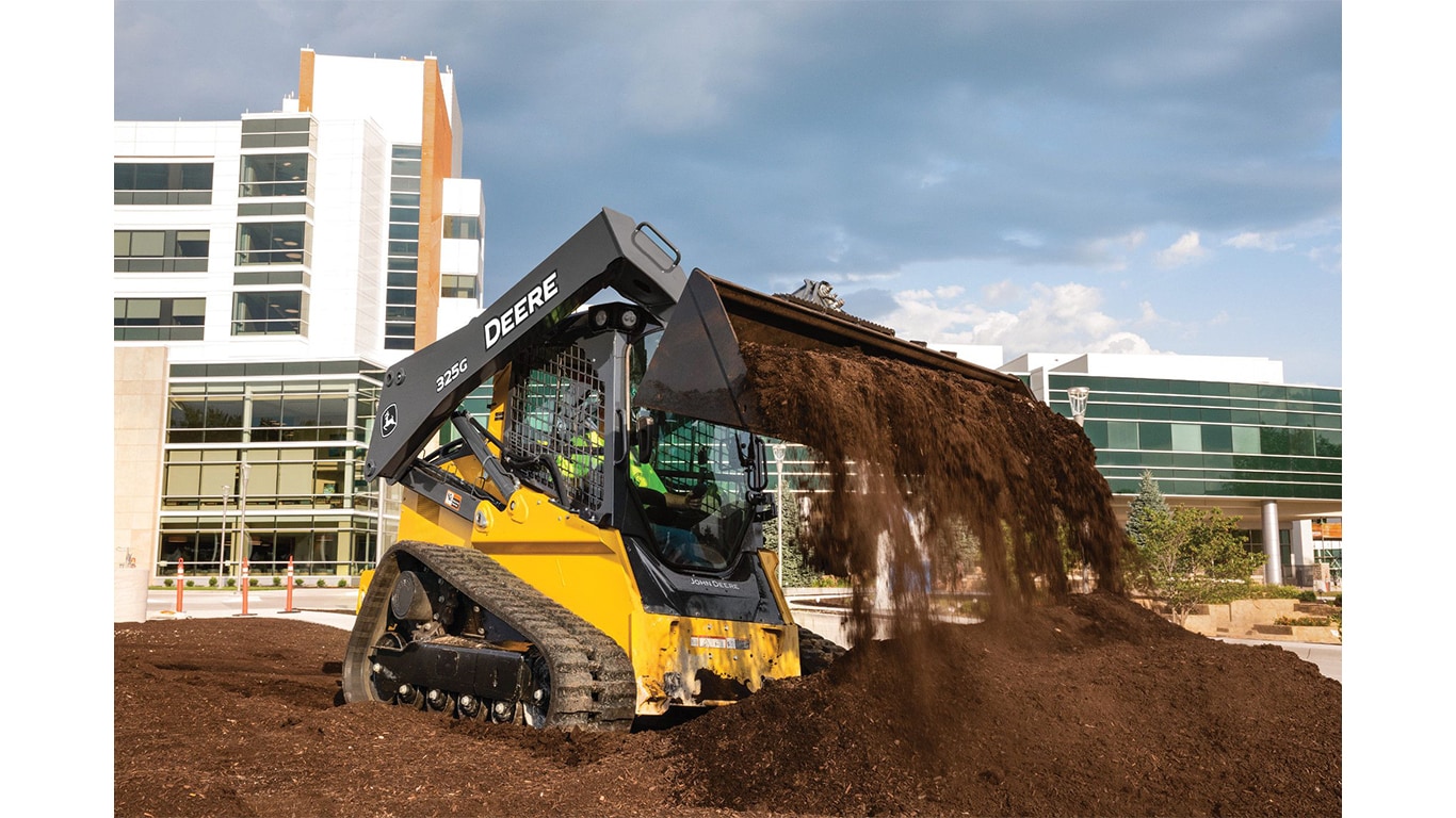 Une chargeuse compacte chenillée 325G qui déplace de la terre sur un chantier avec une construction commerciale en arrière-plan.