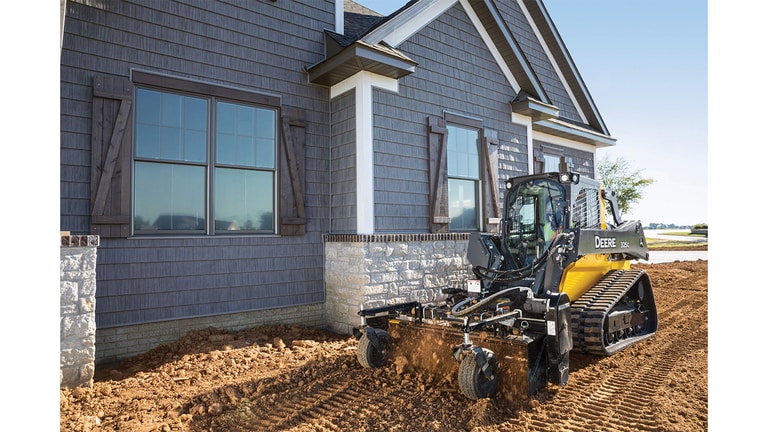 A 325G Compact Track Loader with a power rake attachment raking dirt in front of a house.