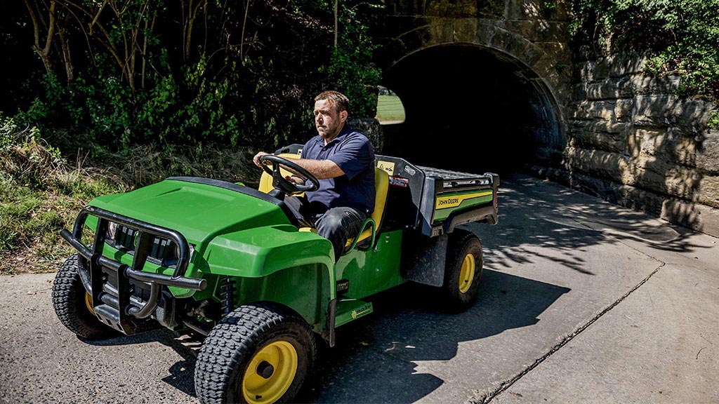 A middle aged man with a brown beard wearing a navy shirt and jeans rides on a green and yellow John Deere electric Gator™ TE 4x2 through a tunnel.