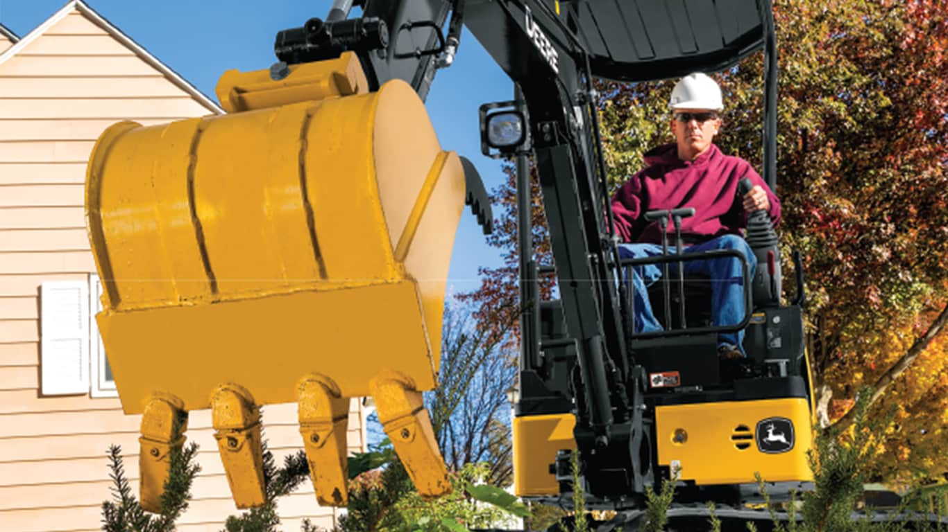 A close up of the 17P-Tier Excavator bucket hovering above plants with an operator and house in the background.