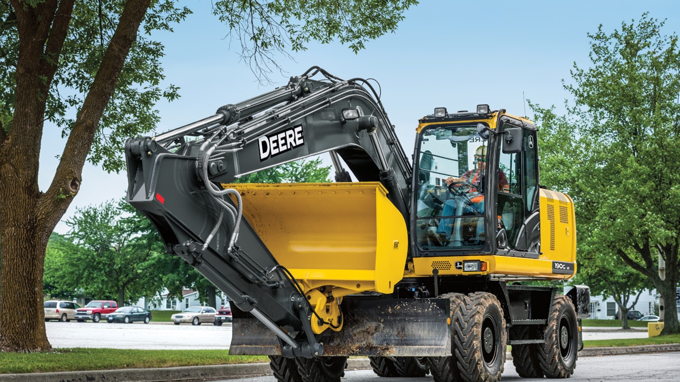 An operator driving a 190GW Excavator down a road with a parking lot in the background.