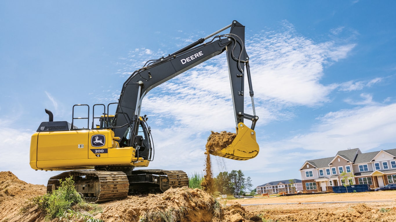 Une excavatrice 200G déplaçant de la terre sur un chantier avec un lotissement en arrière-plan.