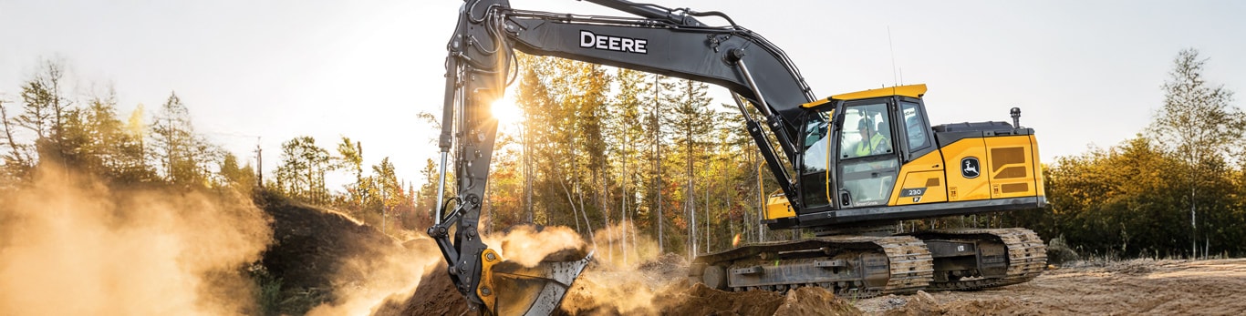 A John Deere 230 P-Tier Excavator is digging a hole with a forest in the background.