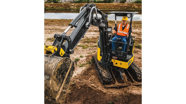 An operator using a 26P-Tier Excavator to scoop dirt at a worksite with a pond in the background.