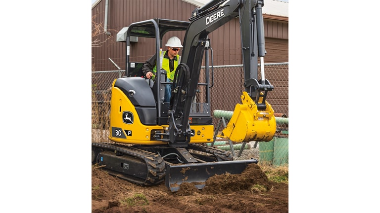 An operator using a 30P-Tier Excavator to move dirt at a worksite with a fence and brown building in the background.