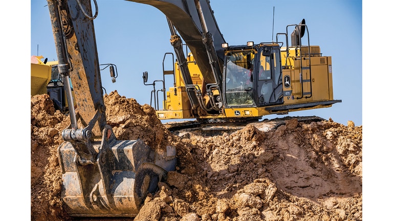 An operator using a 670P-Tier Excavator to scoop up dirt at a worksite with an articulated dump truck to the left.