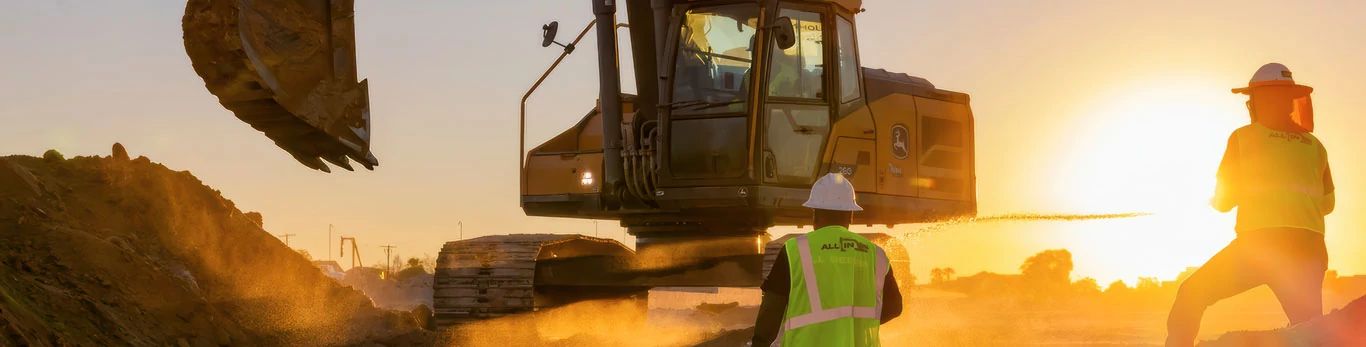 Two construction workers observe John Deere excavator working while the sun rises.