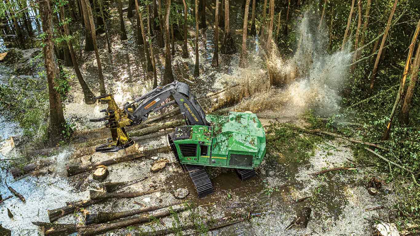 Abatteuse-groupeuse chenill&eacute;e verte qui coupe des arbres dans une for&ecirc;t humide avec de l'eau qui &eacute;clabousse &agrave; proximit&eacute;.