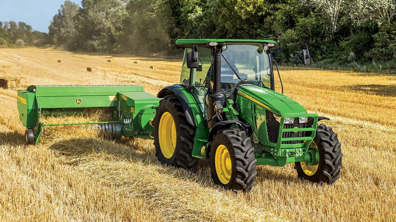 Green tractor with yellow wheels pulls baler through golden wheat field with trees behind.