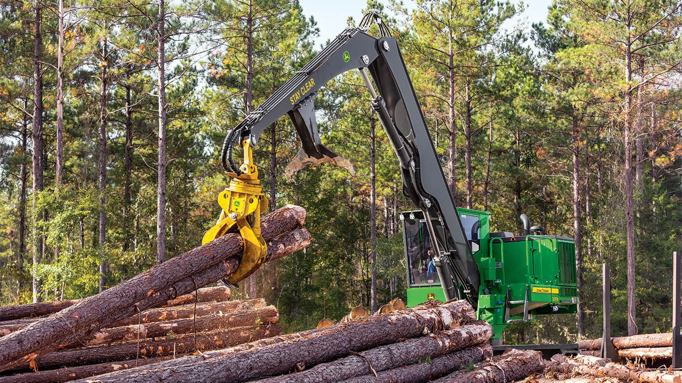 Une grue de chargement John&nbsp;Deere ajoutant des rondins à une pile de rondins dans une forêt.