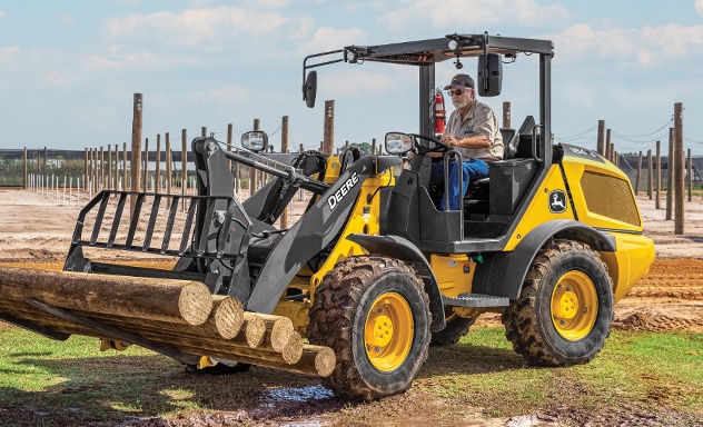 John Deere 184 G-Tier compact wheel loader with fork attachment lifting logs.