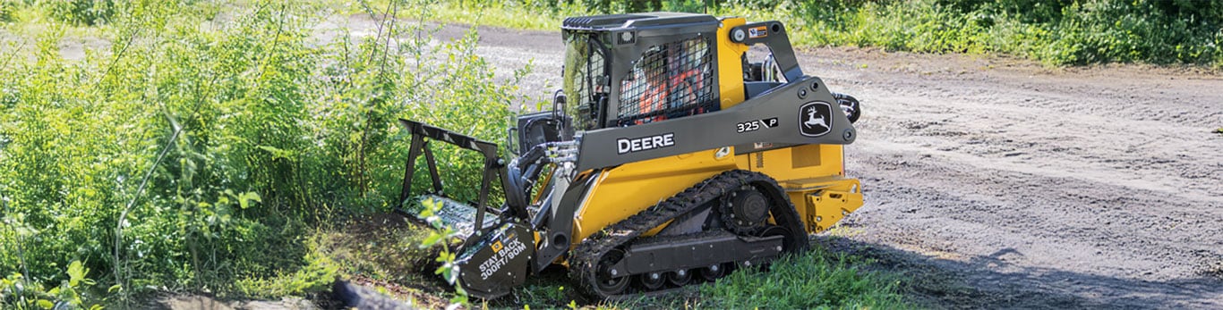 skid steer loader working on a grassy area, surrounded by green vegetation, on a gravel path.