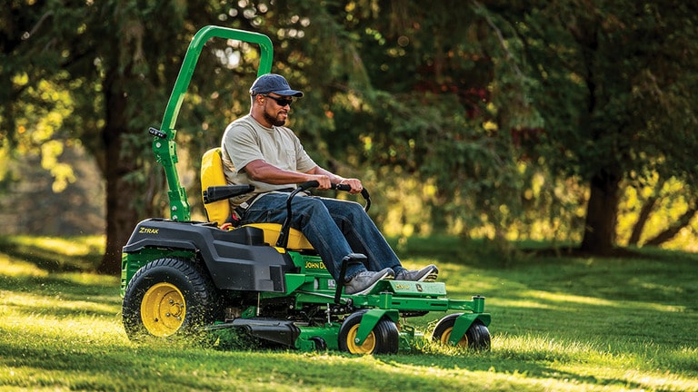 A person with sunglasses and a hat cuts grass with a green zero-turn mower in front of a line of trees.
