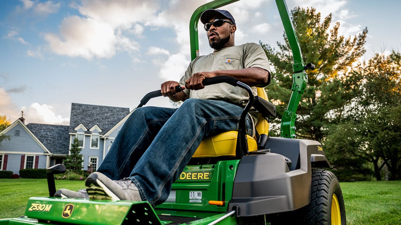 Close-up of a green zero-turn mower being driven with with a large lawn and blue residence in the background.