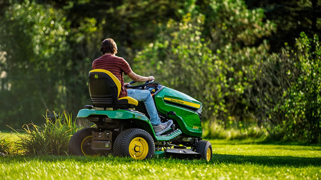 A person driving a green riding mower cutting the lawn around some tall decorative grass.