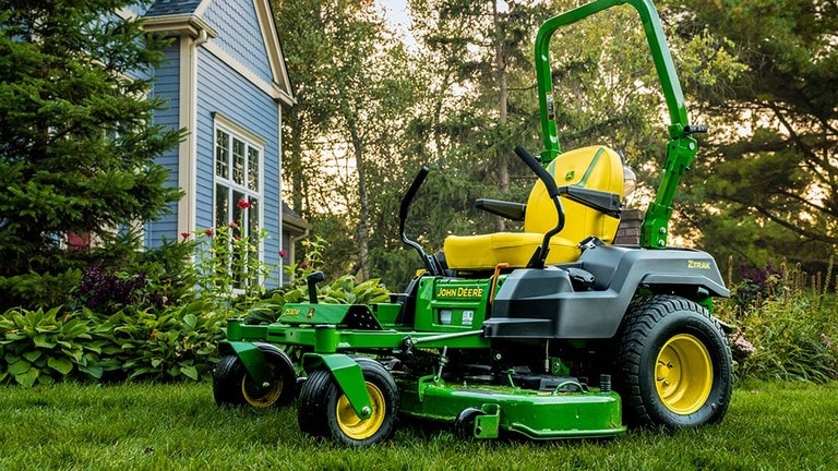 A green zero-turn mower rests on a well-kept lawn near a blue residence and garden.