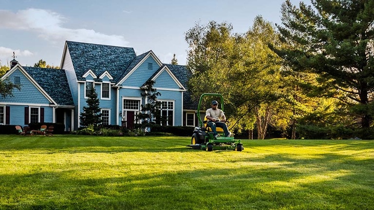 A far-off shot of a green zero-turn mower cutting a spacious lawn behind a large blue house.