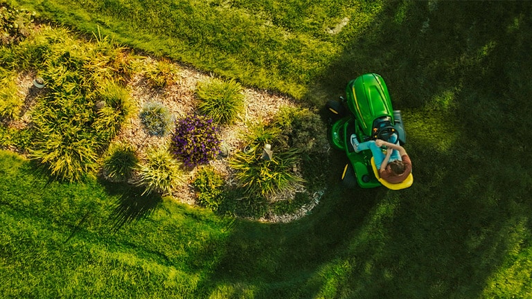 Top-down view of a green riding mower cutting grass around an oval-shaped garden patch.