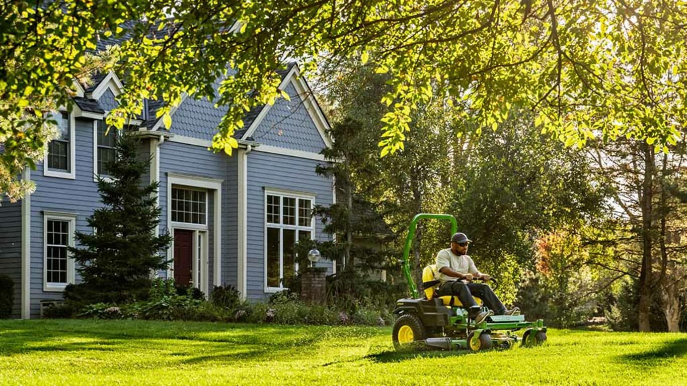 An under-tree shot of green zero-turn mowing along a treeline near a residence on a bright sunny day.